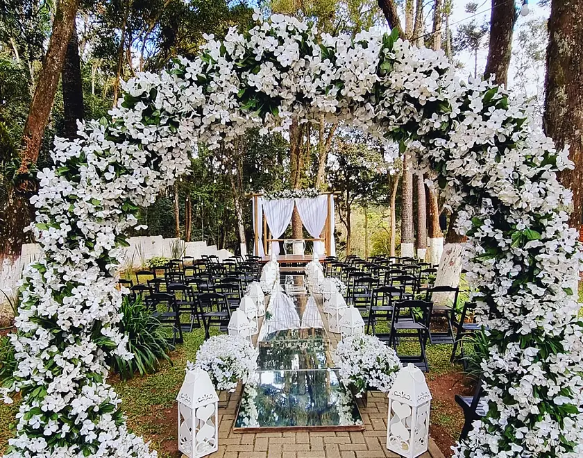 Cerimônia montada no bosque com arco de flores brancas, corredor espelhado e lanternas